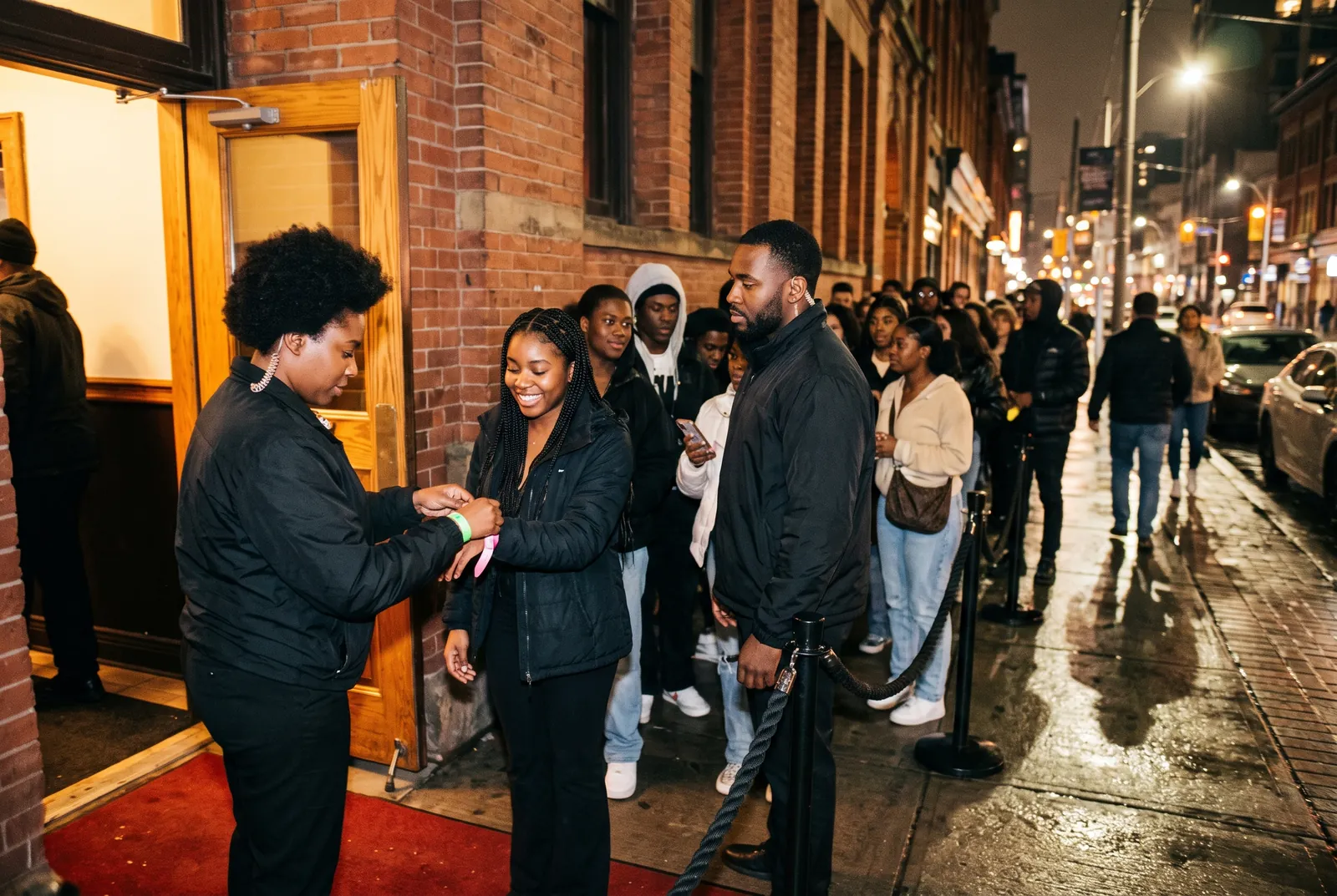 Event security staff check wristbands at a venue door while guests wait in line.